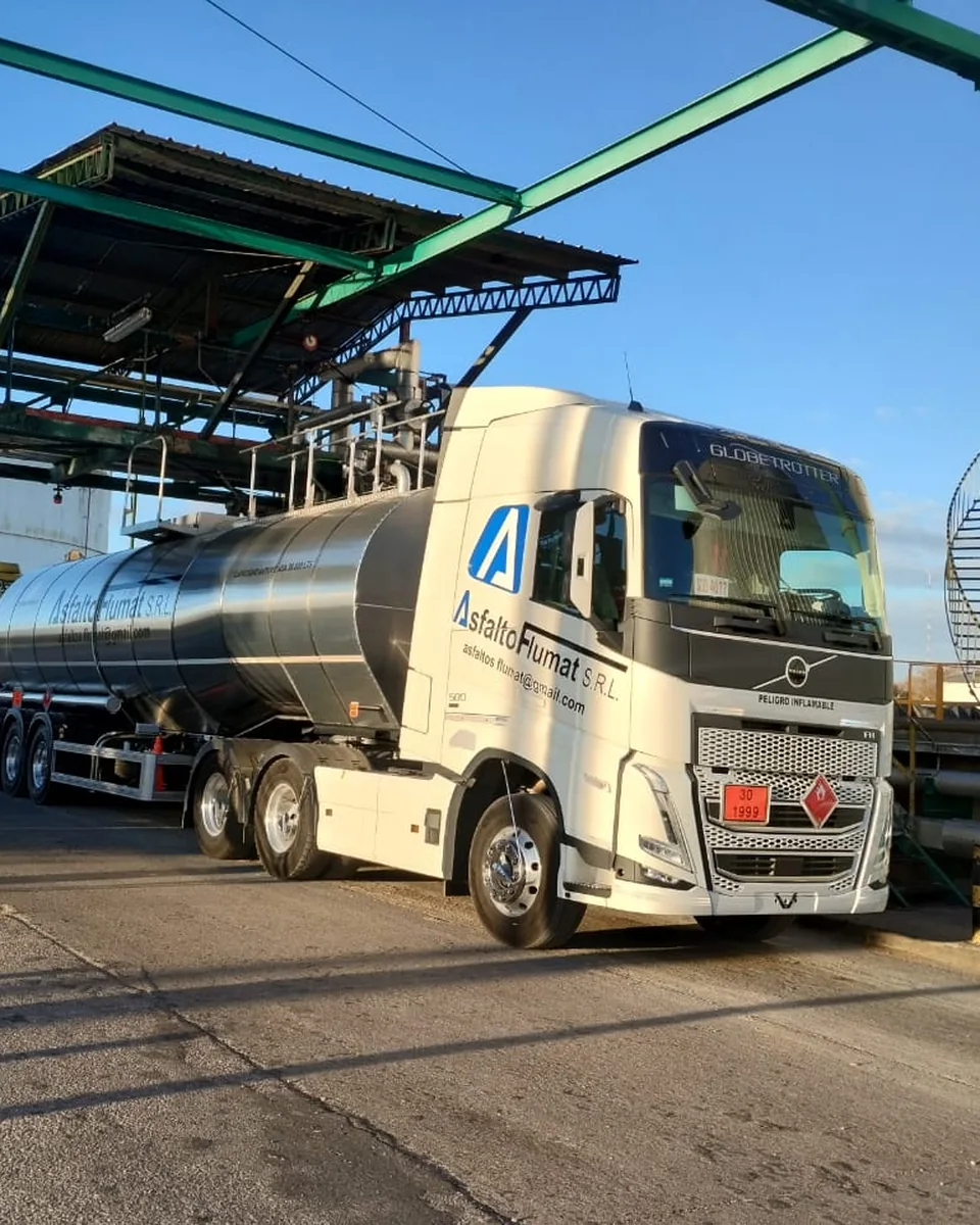 Unidad cisterna de Asfalto Flumat bajo la pasarela de carga de una planta de asfalto: tractor Volvo FH blanco con el logotipo de la empresa y semirremolque cisterna de acero inoxidable pulido, junto al tanque vertical de almacenamiento.