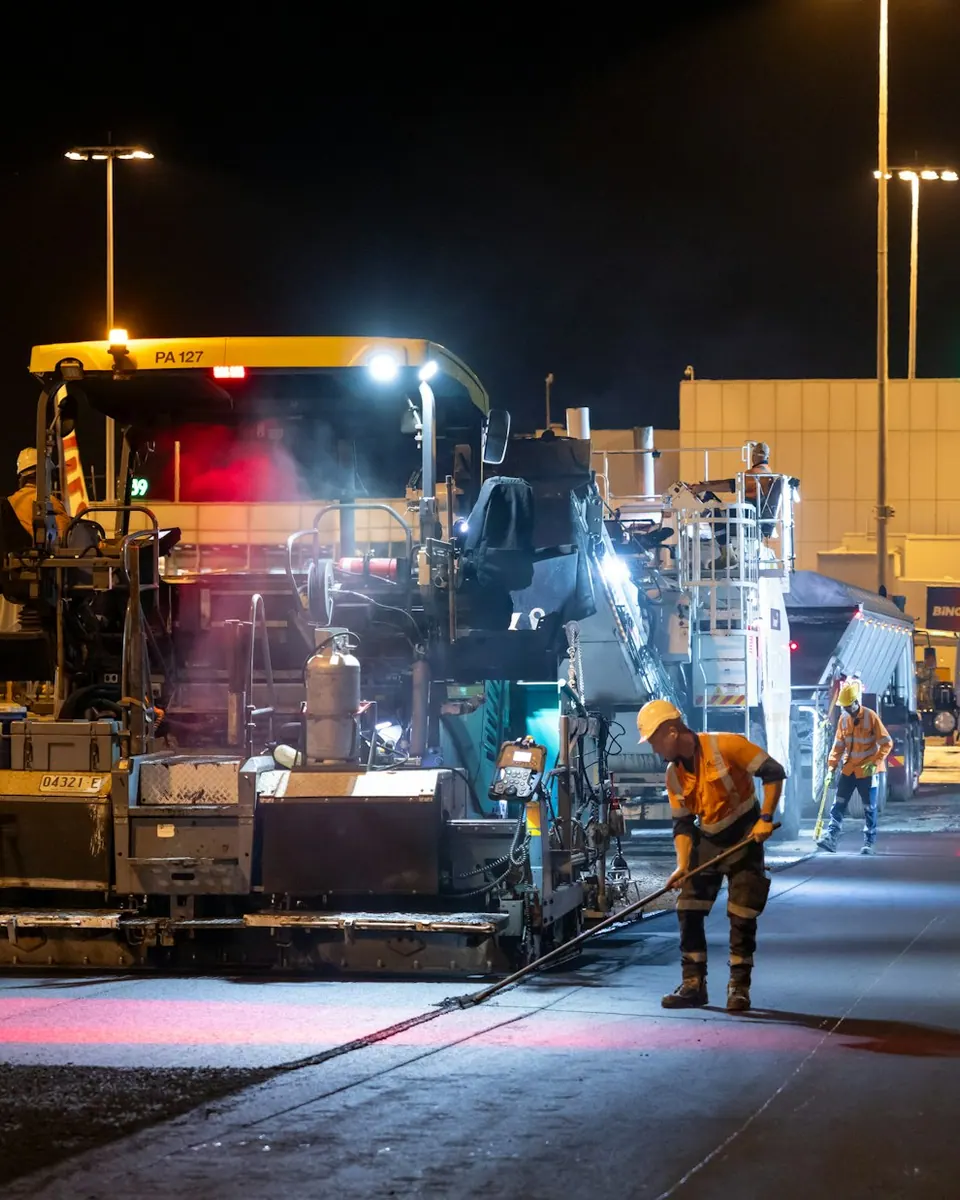 Pavimentación asfáltica nocturna: terminadora iluminada extendiendo carpeta caliente.